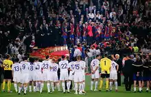 A line of men in white shirts watching men in blue-and-red shirts standing on a rostrum in the background.