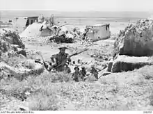 a black and white photograph of helmeted troops carrying rifles with bayonets fixed at the high port climbing up a gully between rocky outcrops with a rustic building in the background
