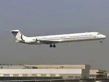 A jet airliner with tail-mounted engines and a T-tail configuration, painted mostly white with a thin red stripe running along the fuselage below the windows, flying low above airport buildings with landing gear and flaps extended.