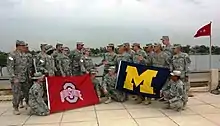 Two groups of soldiers in camouflage uniforms staring at each other on a waterfront; the left group carries a red flag with a silver "O" and the words "OHIO STATE" on it; the right group carries a blue flag with a yellow "M" on it.