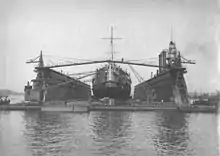 Large ship atop a floating platform with two tall walls running along her side, and construction equipment between the walls and the ship