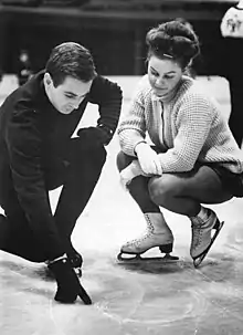 Man on the left and woman on the right crouching on a piece of ice on a rink, the man tracing a figure on the ice with his thumb