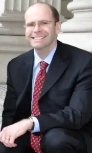A portrait of a bespectacled man wearing a suit, slightly kneeling with his hand on his knee