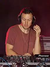 Chris Lake, a white man, stands in front of a soundboard, wearing a red t-shirt and headphones.