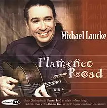 Laucke, seated in half profile, playing his guitar and smiling while looking away from the camera