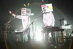 Two people with white comedy space suit costumes ant include a square tv sort of masks on stage in front of instruments and microphones