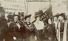 A group of suffragist woman standing together and talking, with banners in the background