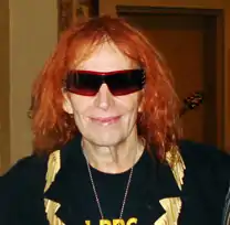 The head and shoulders of a man with ginger hair, wearing sunglasses, a neckless, and a dark jacket with gold trim over the top of a dark T-shirt.