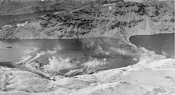 Black and white photograph of a Second World War-era warship in a small bay with a steep and snow-covered shoreline. Clouds of smoke are being blown from the shoreline towards the ship.