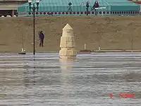 Water from a river completely surrounds a stone obelisk. In the background are several people with news cameras. A building with a green roof is seen behind them. At the bottom right of the image is the camera's time: "6 7:59&nbsp;am".