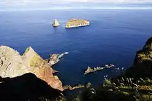Two small volcanic islands with minimal vegetation as they appear from a neighboring island - the ocean, in dark blue, and the sky, in light blue, are also present in this image