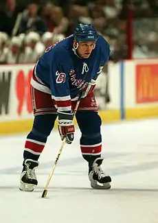 A Caucasian hockey player. He wears a dark blue jersey and helmet. He is bent over at the waist leaning on his stick.