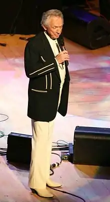 Man in white slacks and black jacket standing singing on stage