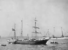 A three-masted ship with sails furled, short funnel amidships, flag flying from the stern on left of picture. Two small boats are close by, and a larger vessel decked with bunting is visible in the background.