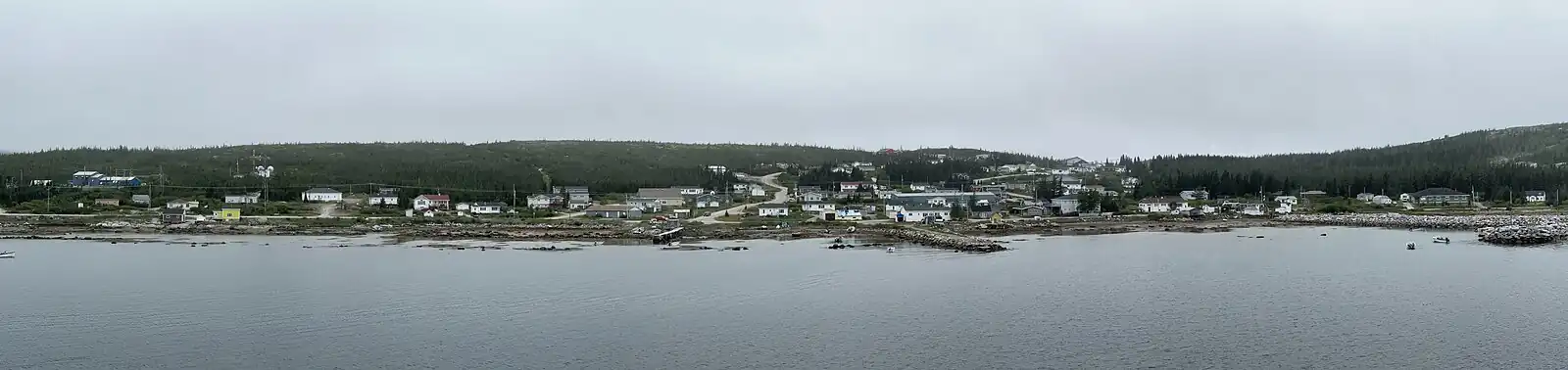 Panorama of small fishing town on a cloudy day with water in foreground, a few dozen buildings, some boats.