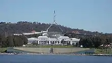 Bright white classical building in front of larger, modernistic beige, gray, and green building. Two metal spires stretch from the top of the larger building in a trangular shape; near the top, before the two connect, they bend straight up, supporting a flagpole and flag.