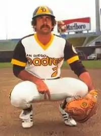 A man in a white baseball uniform crouched down with a catcher's mitt on one hand while giving a pitching sign with the other