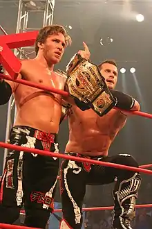 Two male wrestlers standing in a ring holding championship belts wearing black wrestling gear.
