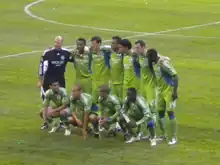 Eleven soccer players wearing green jerseys and green shorts posing in two rows on a grass field.