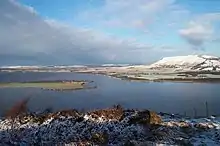 A lake in winter with low snow-covered hills in the background and a brushwood slope in the foreground beneath a mottled blue sky.