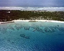 Dark green land with the deep blue sea behind and a lighter blue lagoon in the foreground; the lagoon has isolated reefs visible through its water