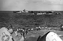 Black and white photo of two aircraft carriers sailing in close formation viewed from the flight deck of another aircraft carrier