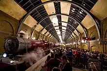 On the left is a red steam locomotive and three carriages, with steam drifting out above. On the right passengers queue behind barriers and signs that say "Hogwarts Express" and "Platform .mw-parser-output .frac{white-space:nowrap}.mw-parser-output .frac .num,.mw-parser-output .frac .den{font-size:80%;line-height:0;vertical-align:super}.mw-parser-output .frac .den{vertical-align:sub}.mw-parser-output .sr-only{border:0;clip:rect(0,0,0,0);clip-path:polygon(0px 0px,0px 0px,0px 0px);height:1px;margin:-1px;overflow:hidden;padding:0;position:absolute;width:1px}9+3⁄4". Over both of these is a large cylindrical glass and steel roof.