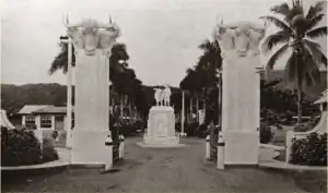 Two white pillars are topped with sculptures of bull's heads. A statue of a man holding a domestic animal can be seen beyond the pillars.