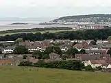 Multiple houses and other buildings around a bay into which a pier projects. On the background are hills.